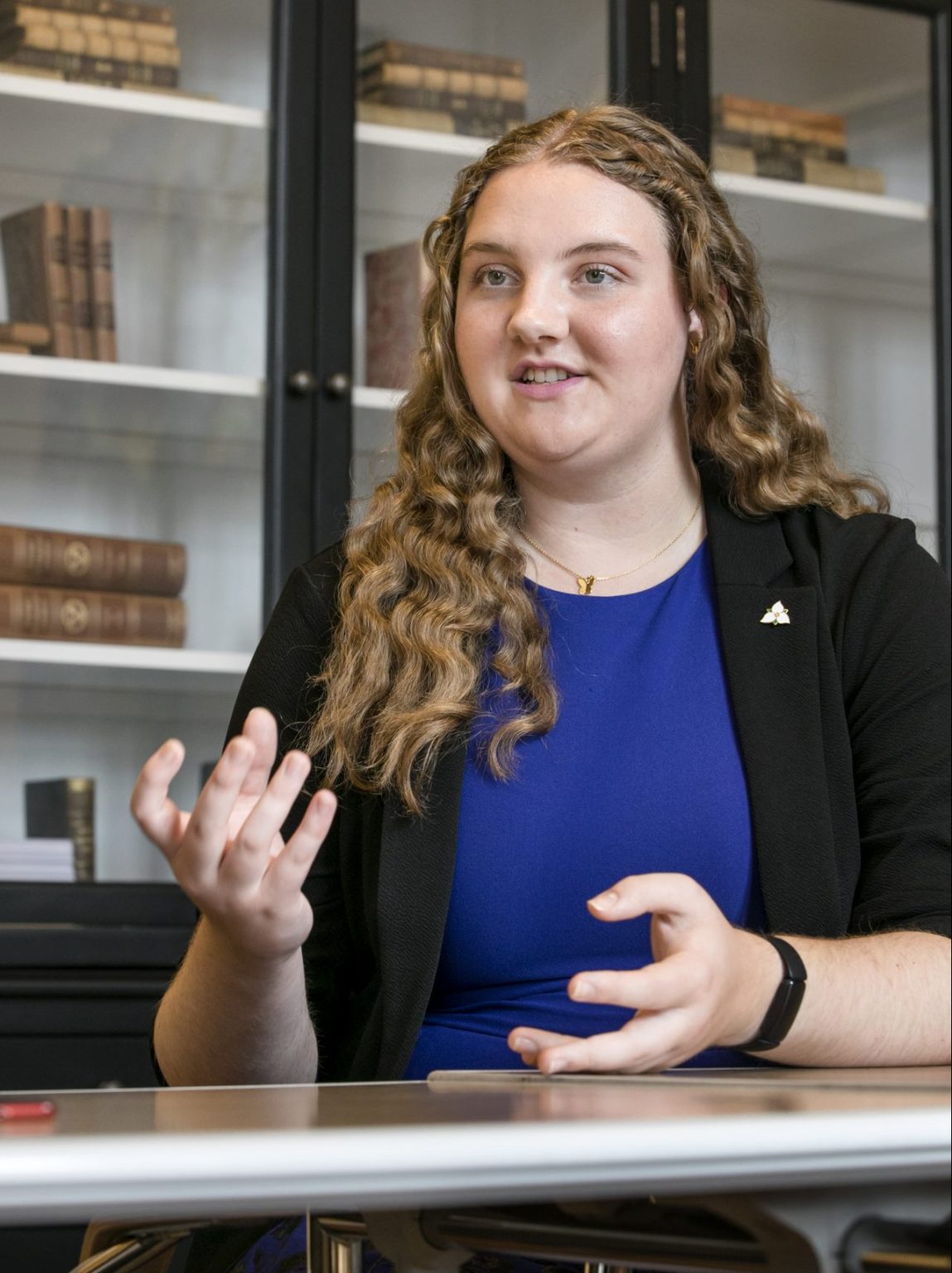 Annabelle looks cheerful while talking, in a black blazer over a blue dress, and long curly hair